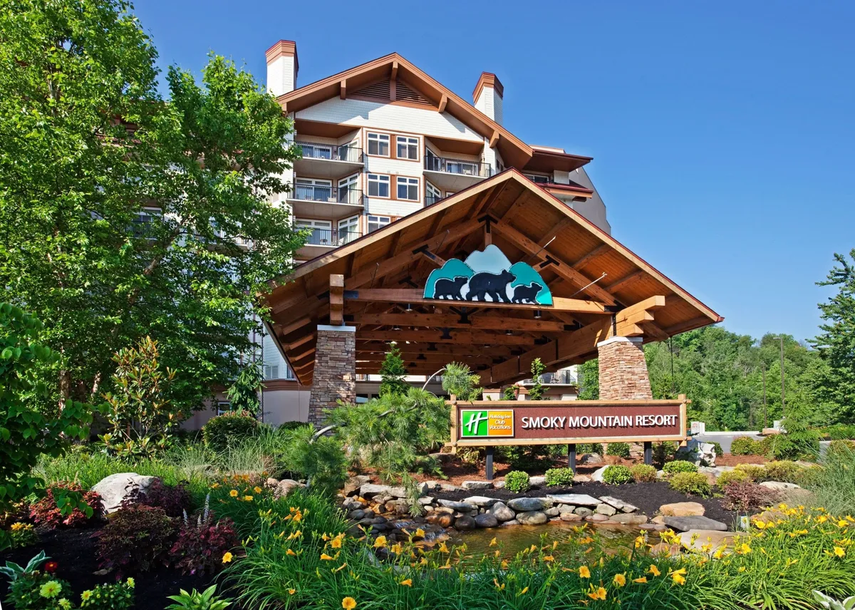 This exterior image showcases the inviting entrance of the Holiday Inn Club Vacations Smoky Mountain Resort. The large wooden canopy creates a grand and welcoming first impression, while lush landscaping and a tranquil water feature set a relaxing, nature-inspired tone. The hotel sign is visible, instantly orienting guests, and people are seen enjoying the outdoors, suggesting a family-friendly atmosphere.
