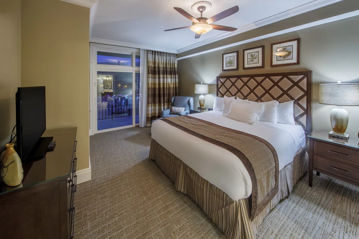 A bright, spacious villa bedroom with a king-sized bed, decorative headboard, nightstands with lamps, a large window with drapes, and a television. The atmosphere is tranquil and restful, ideal for unwinding after a day of exploration.