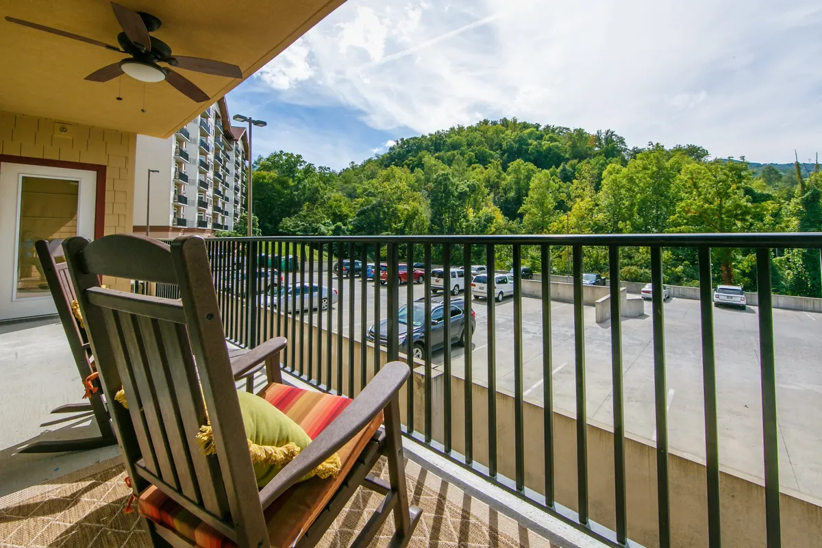An exterior scene showing a relaxing porch area with a classic rocking chair overlooking a lush green hillside. The image also features a visible parking area, fan, and a bright blue sky in the background, creating a welcoming and tranquil outdoor setting.