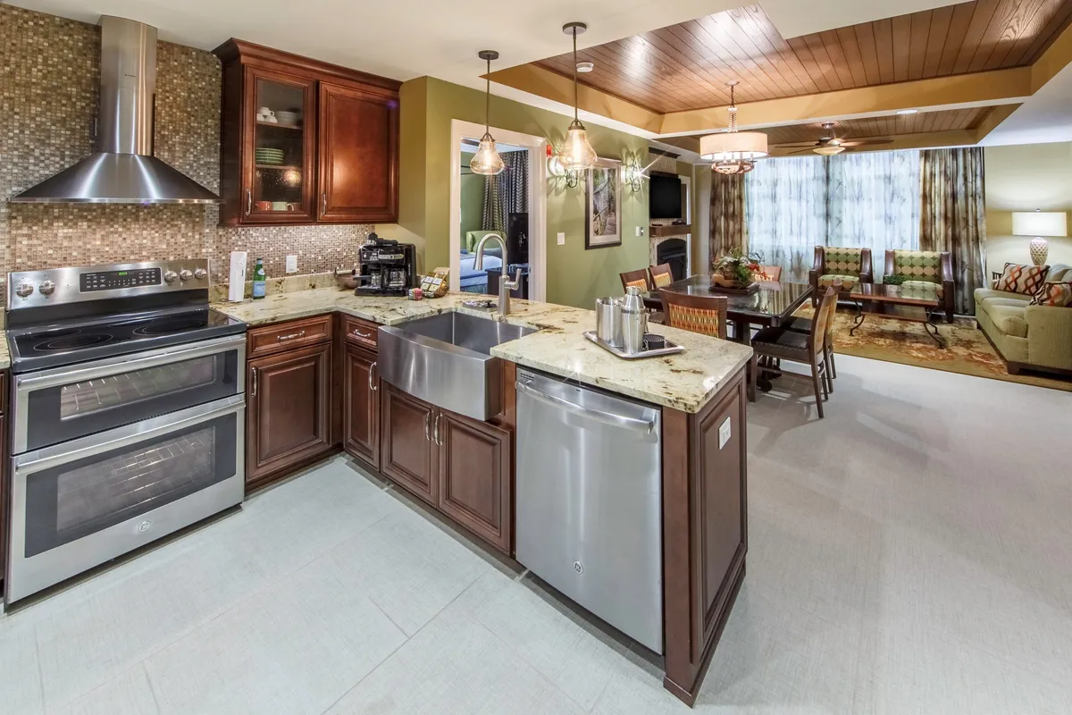 This image captures an open-concept kitchen with a spacious island, stainless steel appliances, and warm wooden cabinetry. The kitchen flows seamlessly into a dining area, which is bathed in natural light from large windows. The overall atmosphere is lively, with hints of holiday feasts or group entertaining in the air.