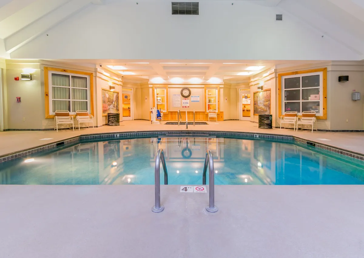A welcoming indoor pool area with tranquil blue water, comfortable lounge chairs, a wall-mounted clock, and a reception area nearby. The atmosphere is relaxed, encouraging both swimming and lounging.