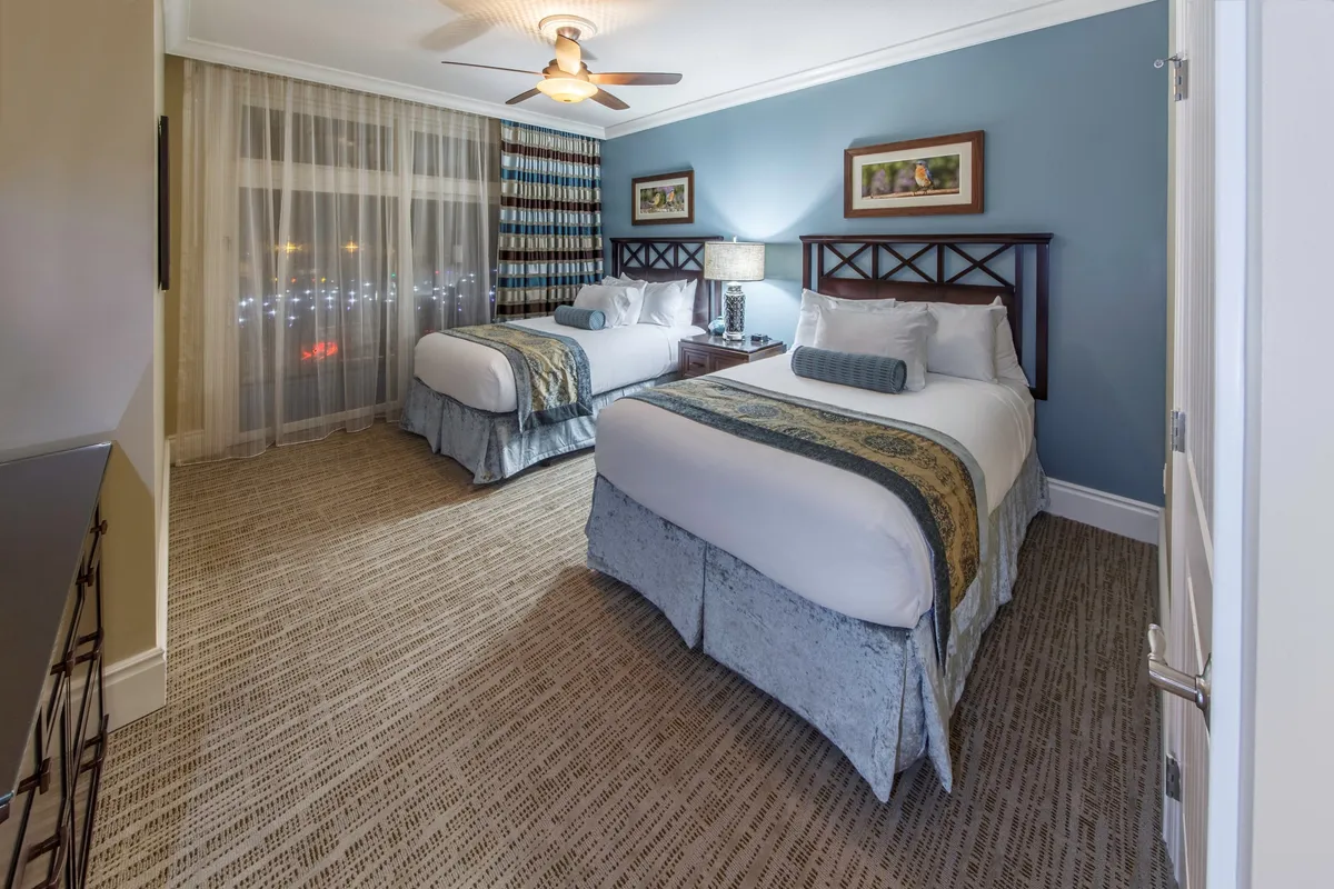 A cozy bedroom set-up featuring two beds with decorative pillows, a central nightstand and lamp, and tasteful framed art for a welcoming ambiance. The space is inviting, ideal for family members or friends sharing a room and creating shared memories.